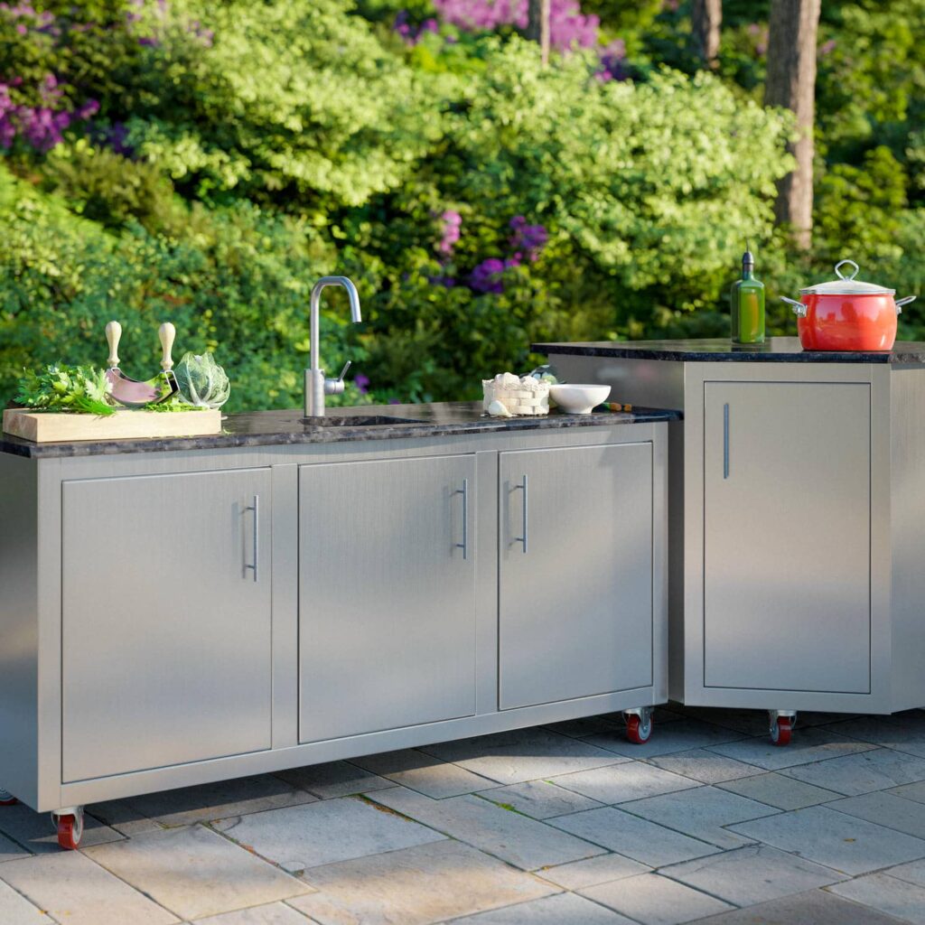 Stainless steel mobile outdoor kitchen with built-in sink and granite prep counter rolling on red casters in a garden setting.