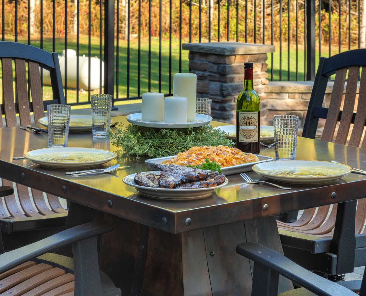 Outdoor patio dining table set with grilled meat, side dishes, drinks, and place settings for a backyard meal.