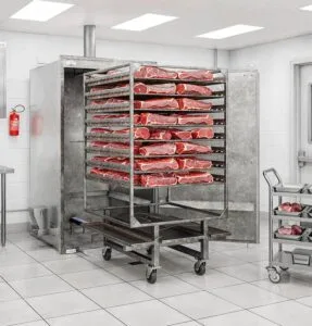 Rocket Outdoors roll-in gas smoker loaded with a rolling rack of raw pork cuts in a commercial kitchen, with a secondary meat cart nearby.