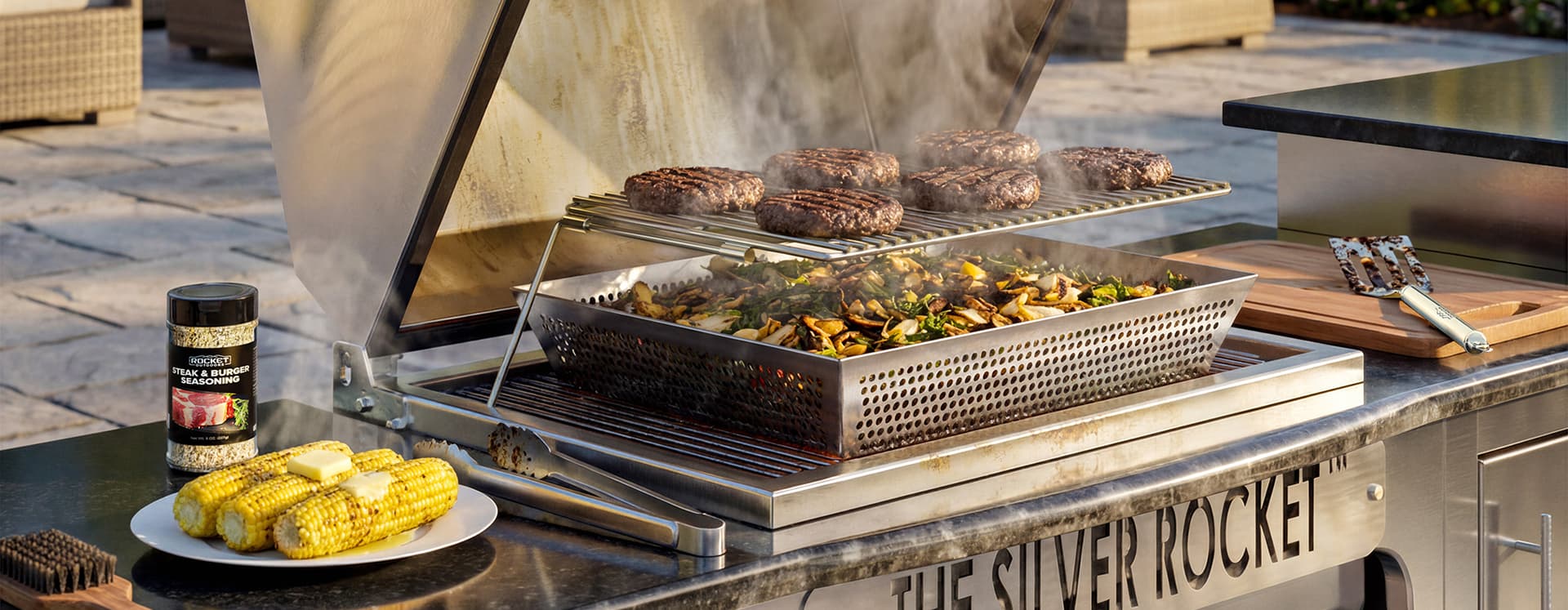 Silver Rocket built-in stainless steel grill cooking burgers and vegetables with corn and steak seasoning on the outdoor kitchen counter.