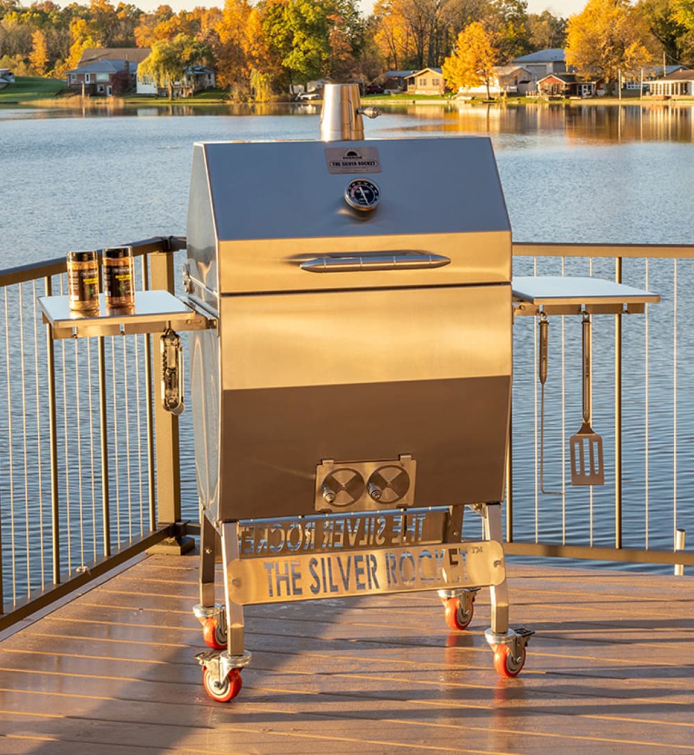 Silver Rocket stainless steel grill on a lakeside deck with side shelves, grilling tools, and a sunset water view.