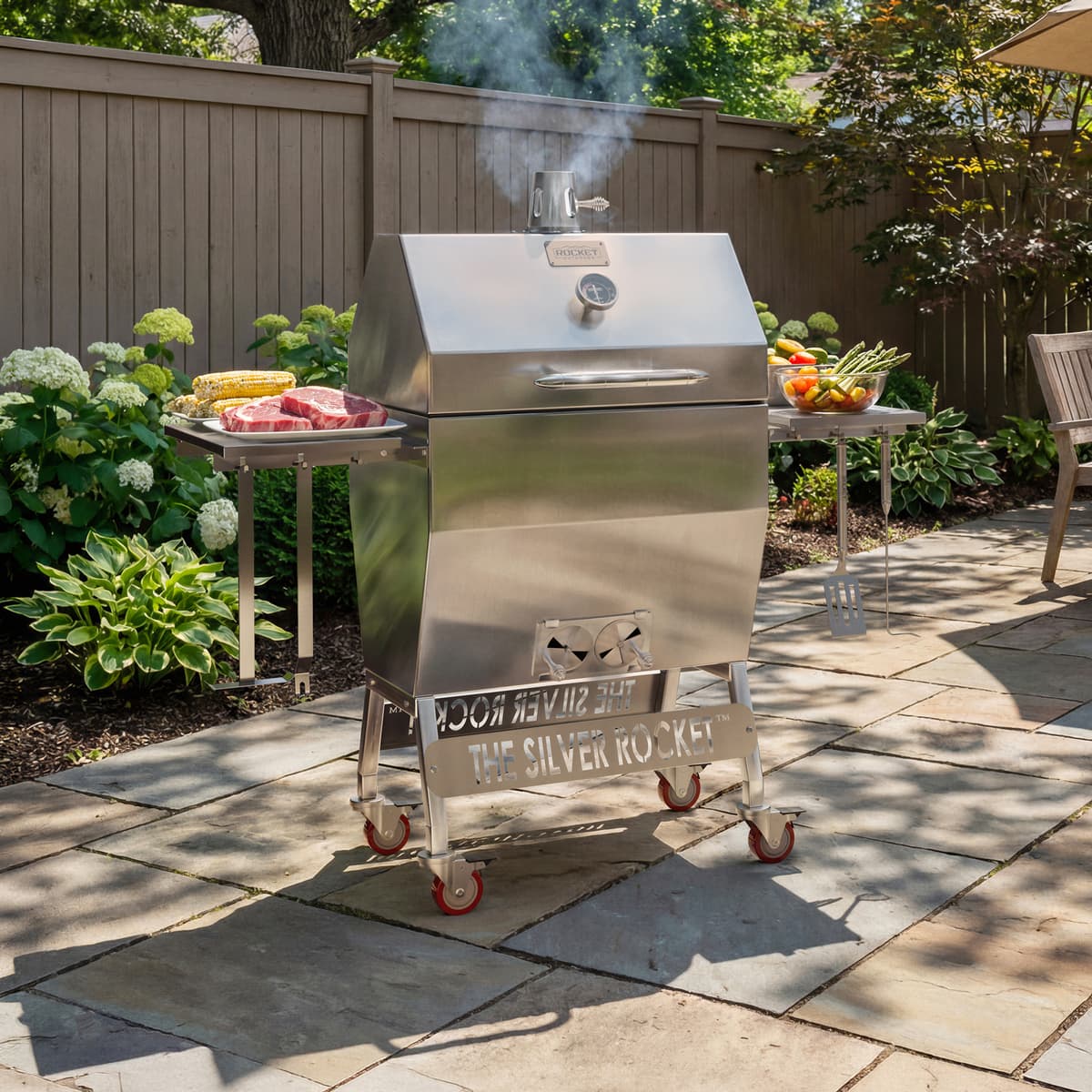 Medium Silver Rocket stainless steel grill on a backyard patio with side shelves, food prep, and smoke coming from the chimney.