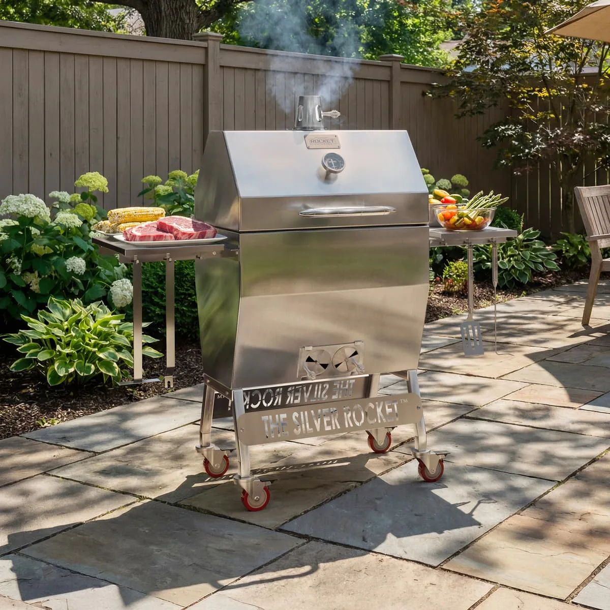 Medium Silver Rocket stainless steel grill on a backyard patio with side shelves, food prep, and smoke coming from the chimney.