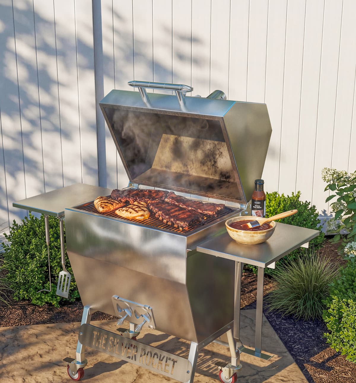 Medium Silver Rocket stainless steel grill with the lid open, cooking ribs and chicken with barbecue sauce on the side shelf.
