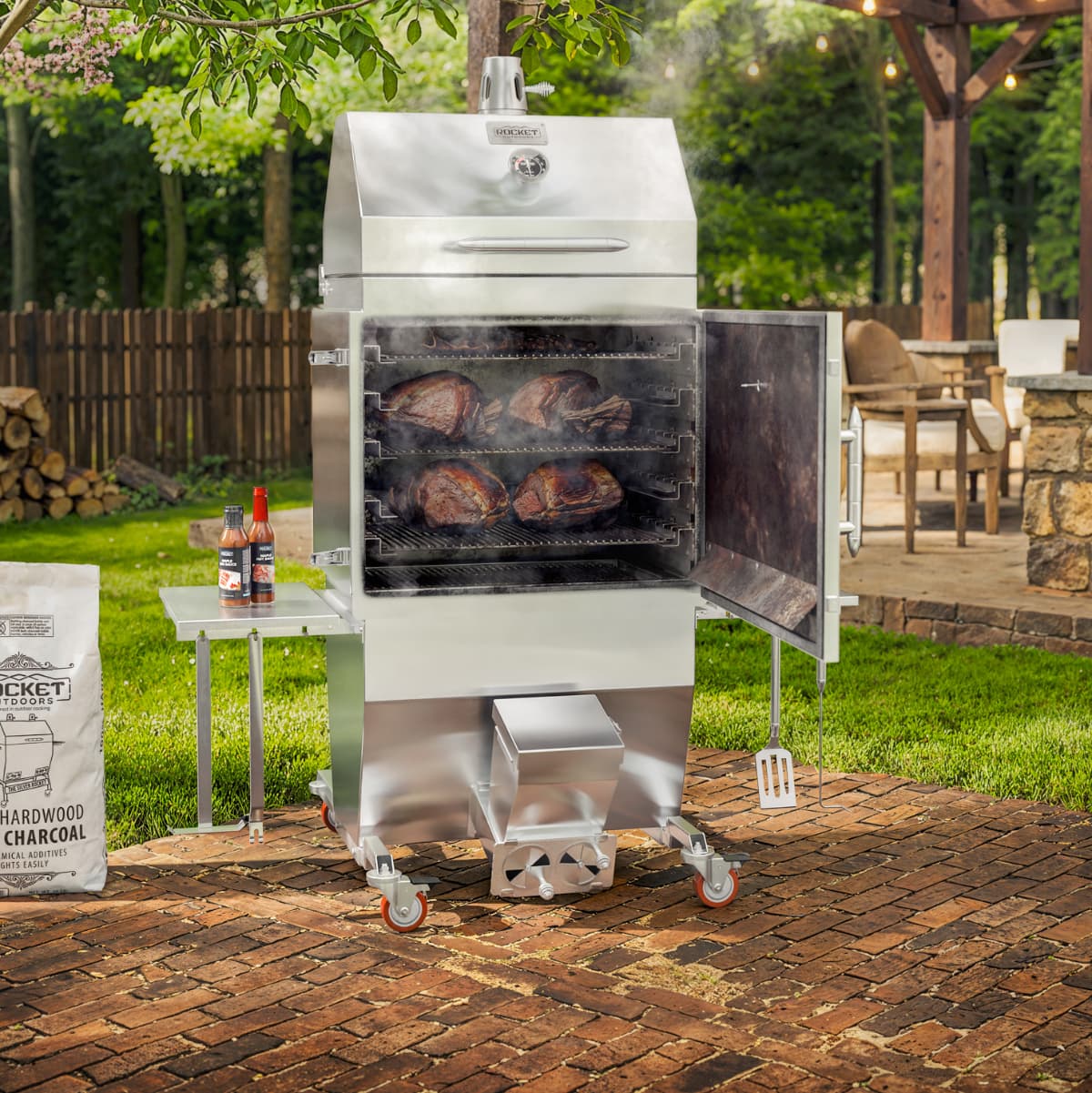 Rocket Outdoors stainless steel smoker baker grill with the door open, showing meat cooking on multiple racks over charcoal.