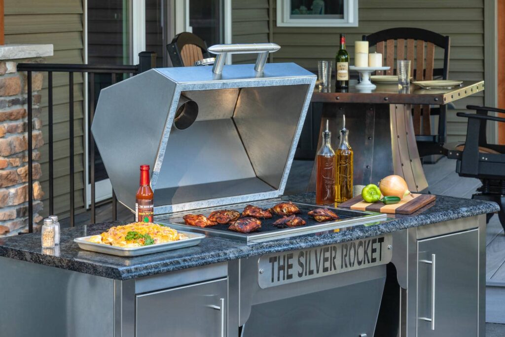 Open Silver Rocket grill with barbecue chicken, a tray of mac and cheese, hot sauce, and fresh produce arranged on the granite countertop of a covered deck.