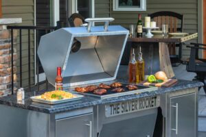 Open Silver Rocket grill with barbecue chicken, a tray of mac and cheese, hot sauce, and fresh produce arranged on the granite countertop of a covered deck.
