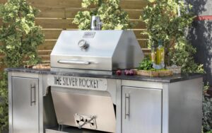 Closed Silver Rocket grill cabinet with fresh herbs, red onions, and olive oil arranged on the granite countertop against a wooden fence.