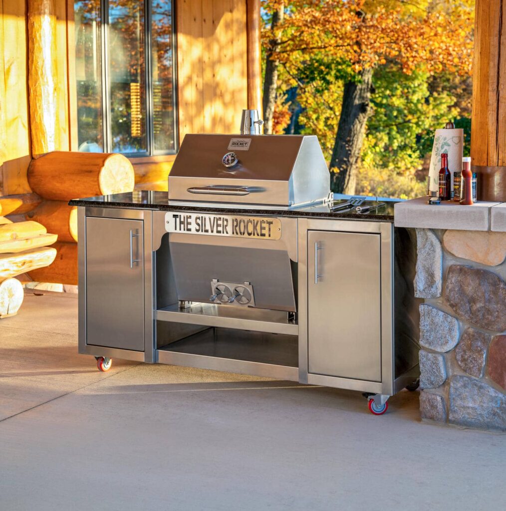 Silver Rocket stainless-steel grill cart with granite countertop on a covered log cabin patio with autumn foliage in the background.