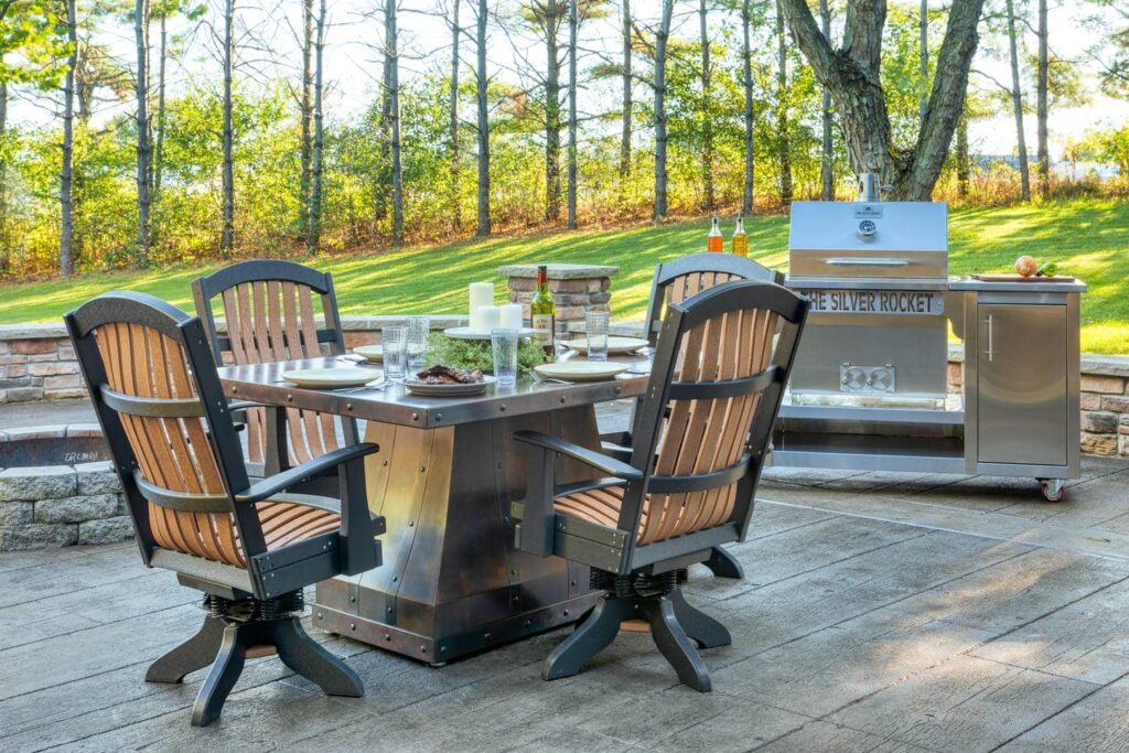 The Silver Rocket grill cart beside a metal-top patio dining table and matching swivel chairs in a wooded backyard setting.