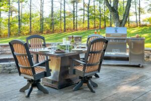 The Silver Rocket grill cart beside a metal-top patio dining table and matching swivel chairs in a wooded backyard setting.