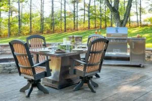 The Silver Rocket grill cart beside a metal-top patio dining table and matching swivel chairs in a wooded backyard setting.