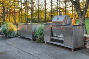 Two-piece Silver Rocket outdoor kitchen with built-in grill cart and prep cabinet rolling on casters across a stone patio in autumn light.