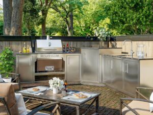 L-shaped Silver Rocket stainless steel outdoor kitchen with built-in grill on a brick patio set with a dining table and pizza.